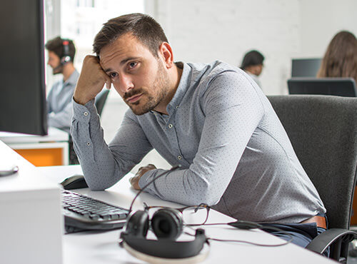Noise and poor acoustics in the office and call centre A man sits stressed and tired at his desk in the office without sound insulation.